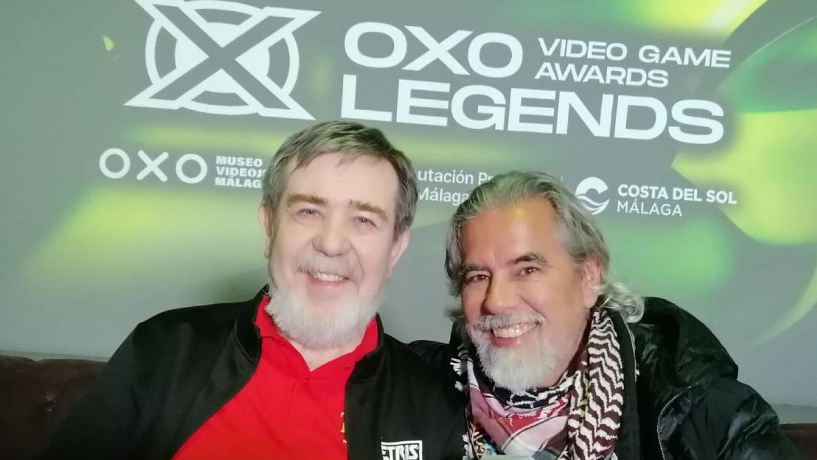 Dos hombres sonríen juntos frente a un panel con logos. Uno de ellos, con camisa roja y chaqueta con el logo de Tetris, posa junto a otro con chaqueta negra y bufanda estampada.