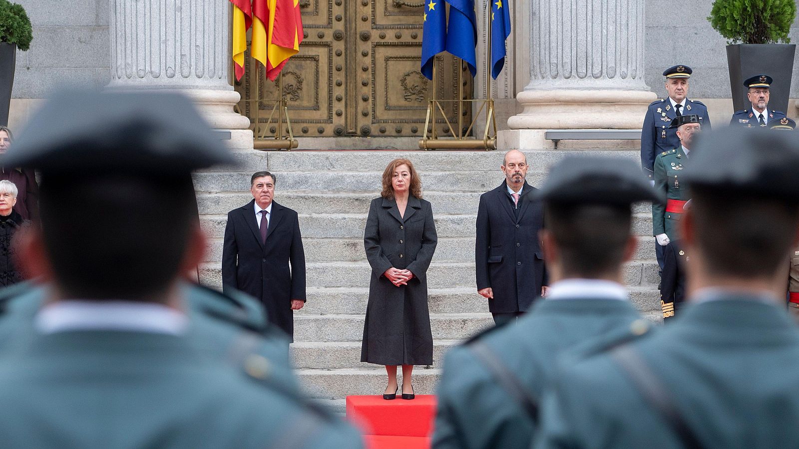 Un acto institucional en España, posiblemente el Día de la Constitución, muestra militares uniformados y una guardia de honor. En el fondo, tres figuras centrales, incluyendo una mujer, observan el evento frente a banderas.