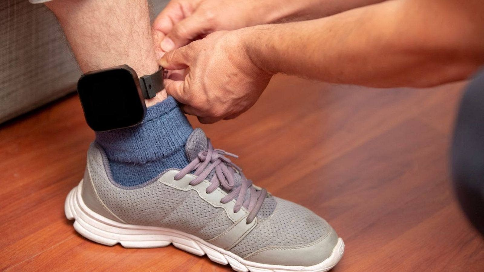 Se coloca una pulsera negra cuadrada en el tobillo de una persona con un calcetín azul y zapatilla gris.  Las manos de otra persona ajustan la correa.
