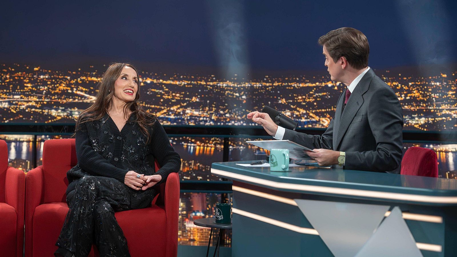En el plató de un programa de televisión, una cantante con conjunto negro brillante conversa con el presentador, quien viste un traje gris y corbata roja, frente a un decorado que simula una ciudad nocturna.