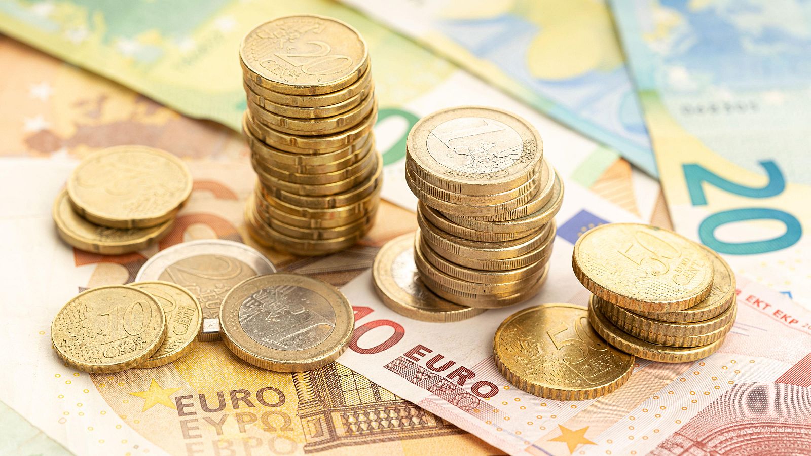 Una fotografía muestra una acumulación de monedas y billetes de euro. Se distinguen dos pilas de monedas doradas, monedas sueltas de diferentes valores y billetes parcialmente visibles.