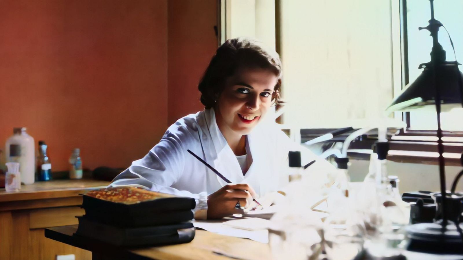 Dorotea Barnés, con bata blanca, está sentada en un laboratorio, frente a una mesa con libros y frascos. Sostiene un pincel o pluma, posiblemente dibujando o escribiendo.