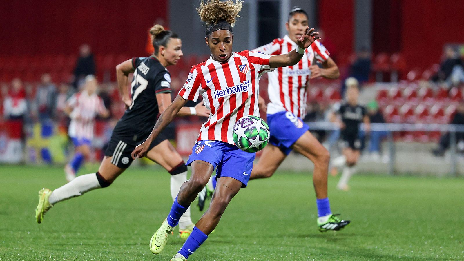 Una jugadora del Atlético de Madrid, con camiseta a rayas y pantalón azul, controla el balón en un partido de fútbol. Se observa a otras jugadoras en el campo, con el patrocinador "Herbalife" en la camiseta de la jugadora.