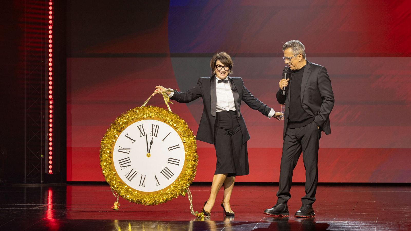 Silvia Abril, con gafas y un traje negro, y Andreu Buenafuente, con un micrófono, comparten un escenario iluminado, posiblemente para un evento especial.