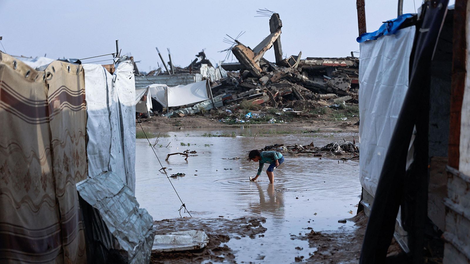 Un niño en un campamento inundado juega en el agua, que le llega hasta las rodillas. El campamento, construido con materiales improvisados, se encuentra en una zona con escombros.