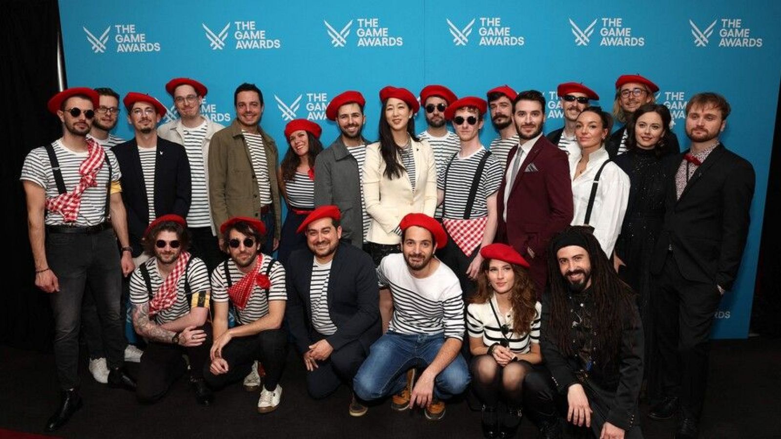 Un equipo numeroso, compuesto por hombres y mujeres, posa para una foto. Visten principalmente boinas rojas y camisetas a rayas horizontales, con algunos accesorios como tirantes y pañuelos a cuadros. El fondo es de color azul claro y muestra un logo repetido.