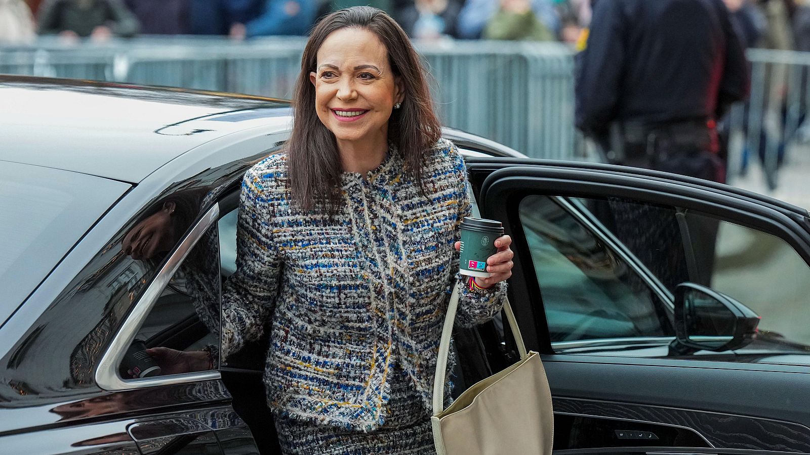 María Corina Machado, con una sonrisa, sale de un coche, vestida con un conjunto de tweed y un bolso beige, portando un vaso de café.