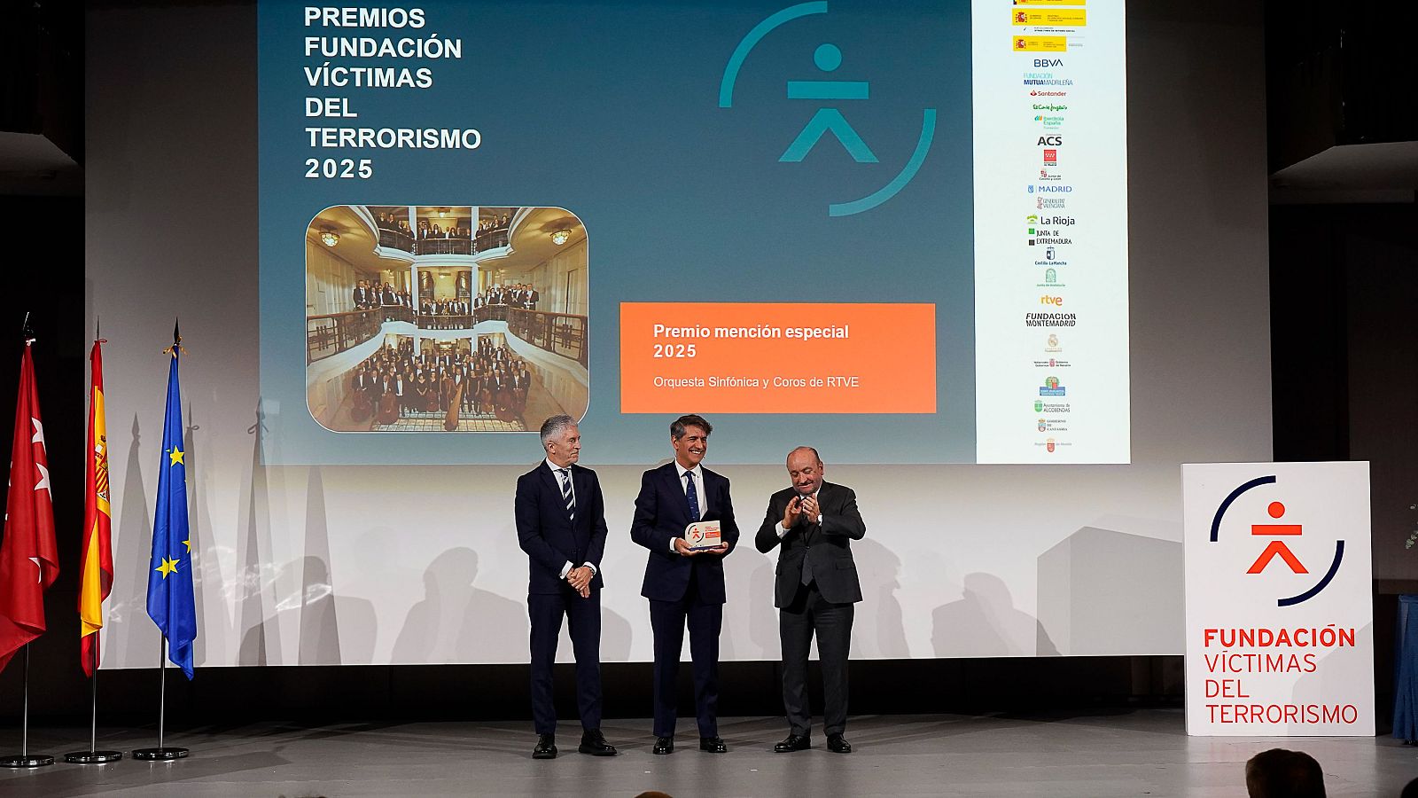 En el escenario de una ceremonia de premios, tres hombres posan frente a una pantalla que anuncia el evento y el reconocimiento a una orquesta y coro. Se observan banderas y logotipos de patrocinadores.