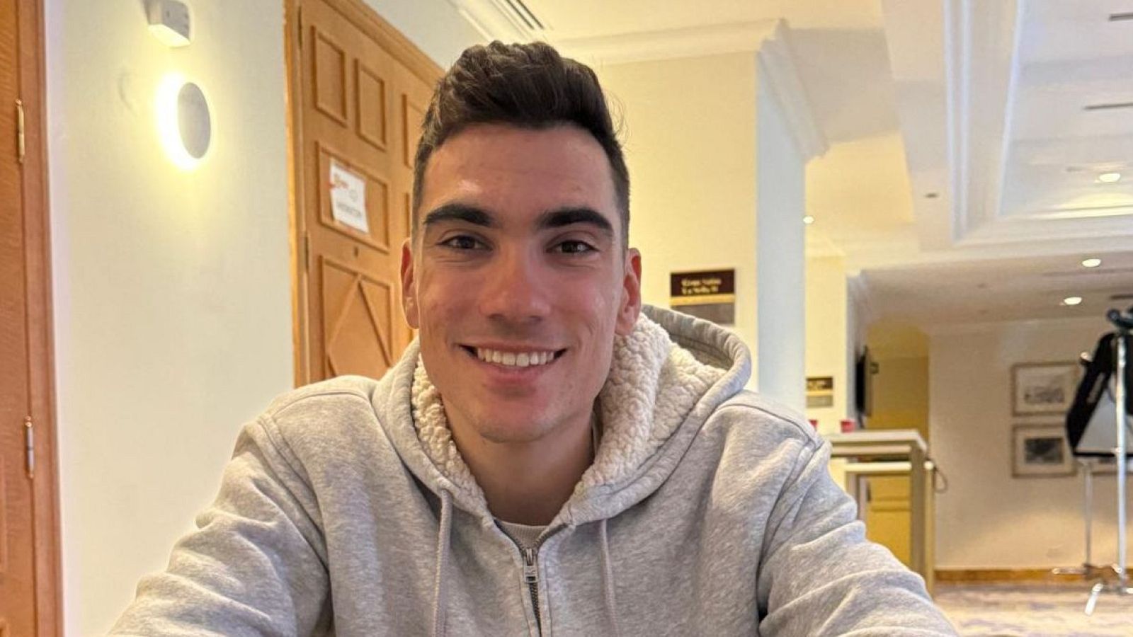 Juan Ayuso, con el pelo castaño oscuro peinado hacia arriba, sonríe mientras viste una sudadera gris. El fondo muestra un interior con una puerta de madera y una mesa con una cámara.