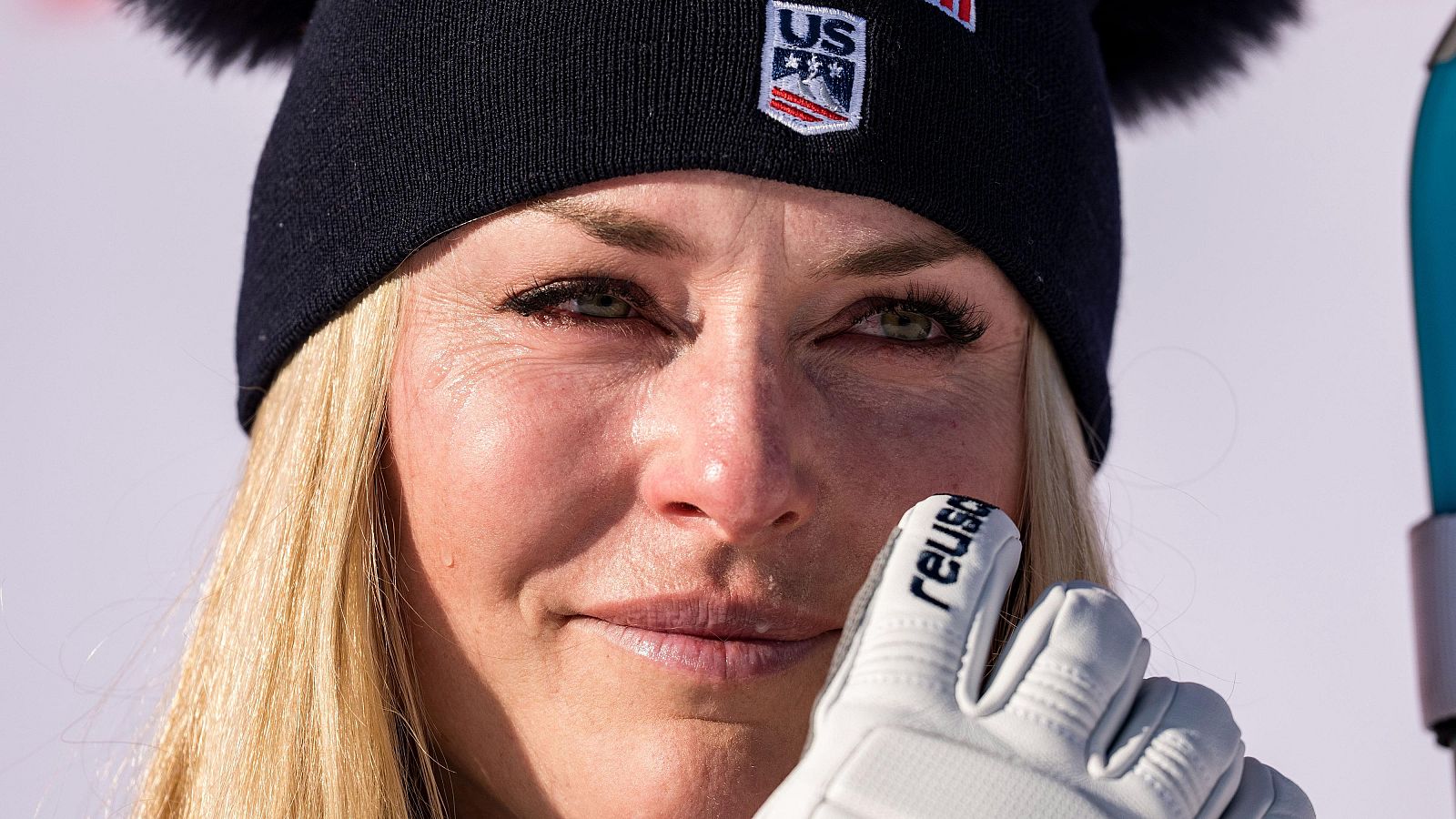 Lindsey Vonn, con cabello rubio y ojos claros, muestra una expresión emocional, posiblemente lágrimas, mientras lleva un gorro azul marino con el logo de un equipo de esquí y un guante blanco.