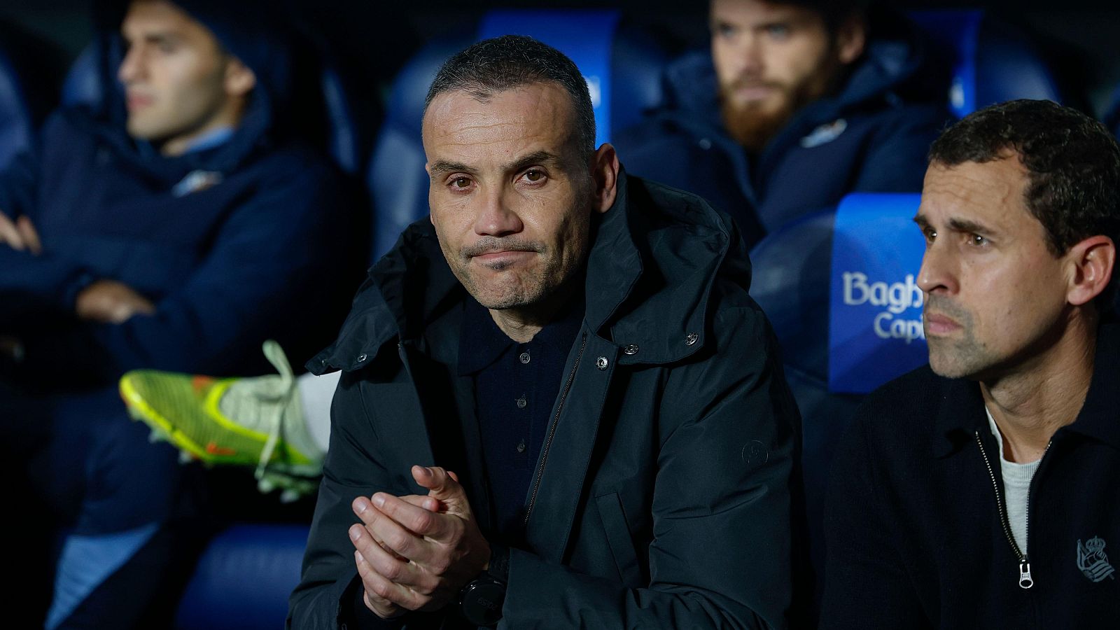 En el banquillo, un hombre con chaqueta oscura y camisa tipo polo oscura está sentado con las manos juntas, acompañado por otra persona con chaqueta oscura. Se distinguen otros miembros del equipo y un logo "Bagh".