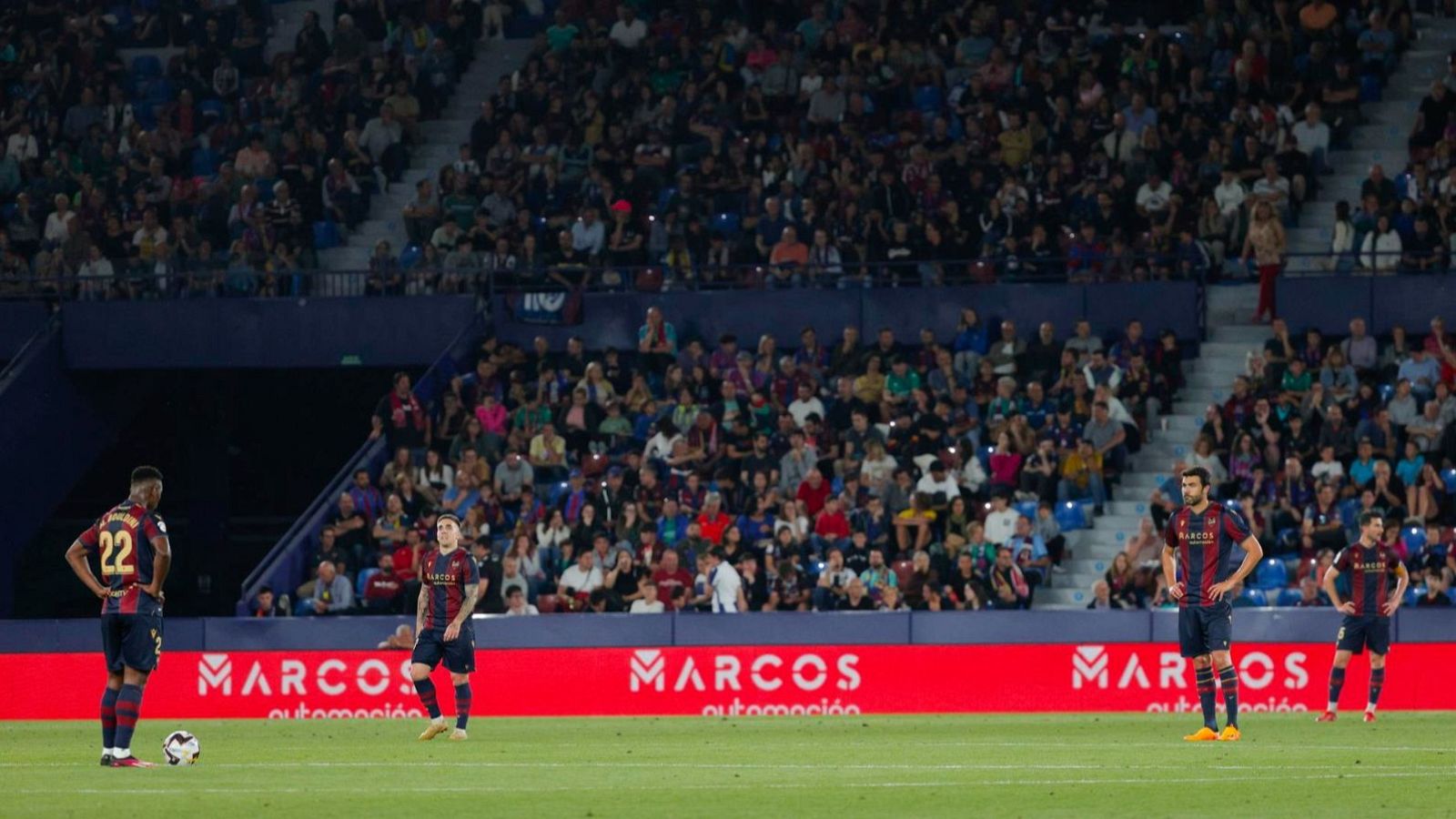En el estadio Ciudad de Valencia, un partido de fútbol muestra a jugadores con equipaciones azulgranas, uno de ellos con el número 22, y un balón en el césped, con una valla publicitaria de fondo.