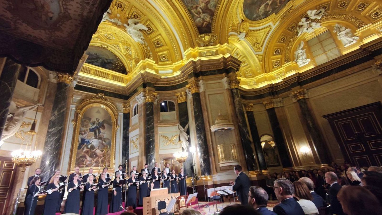 Un concierto navideño de la Orquesta y Coro, con un coro formalmente vestido frente a una audiencia, en un espacio con arquitectura clásica y detalles ornamentales, incluyendo columnas y una lámpara de araña.