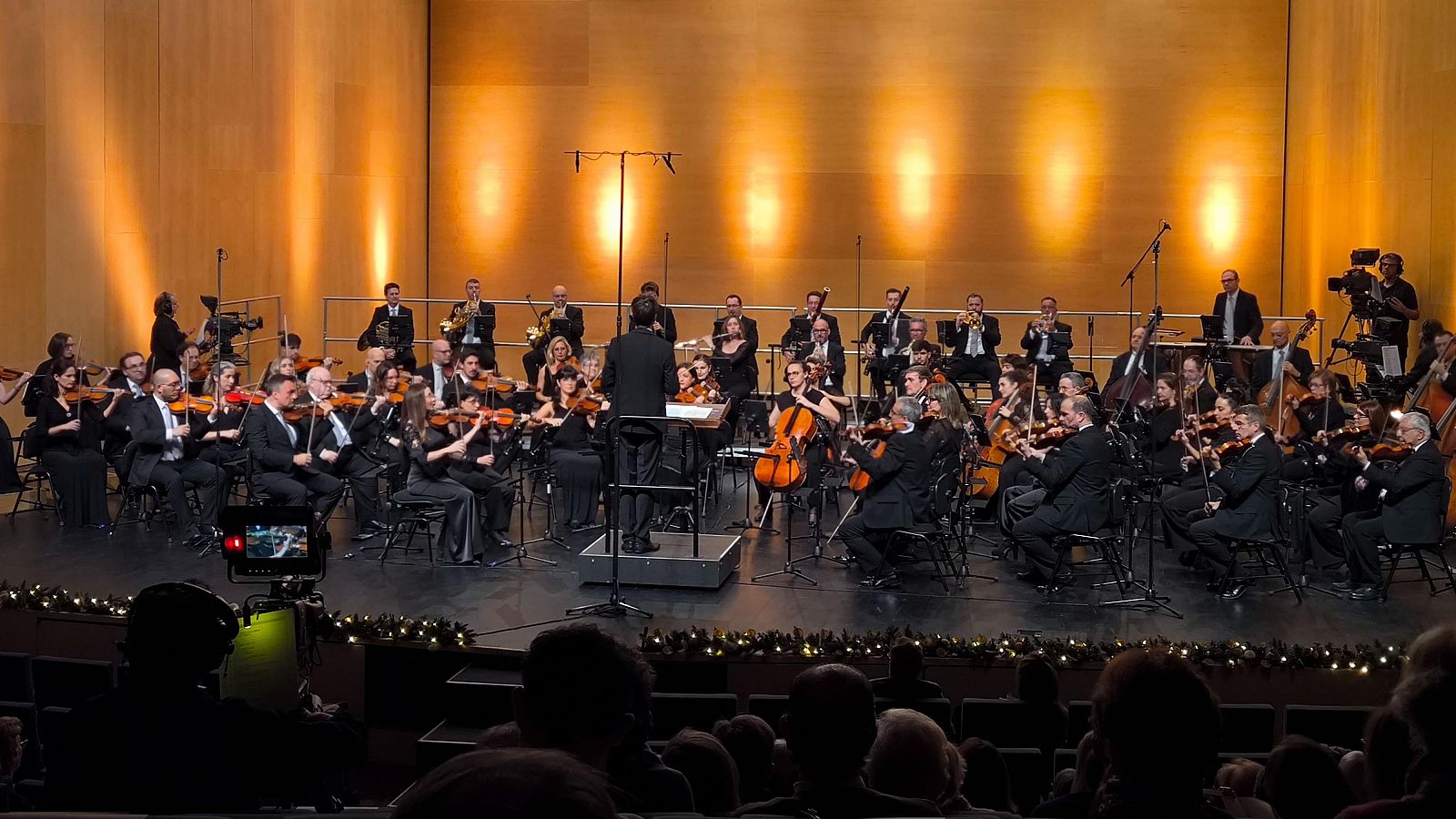 Una orquesta y un coro, vestidos formalmente, actúan en un escenario iluminado, con un director dirigiendo. El público observa la actuación desde las butacas del auditorio, mientras una cámara graba el evento.