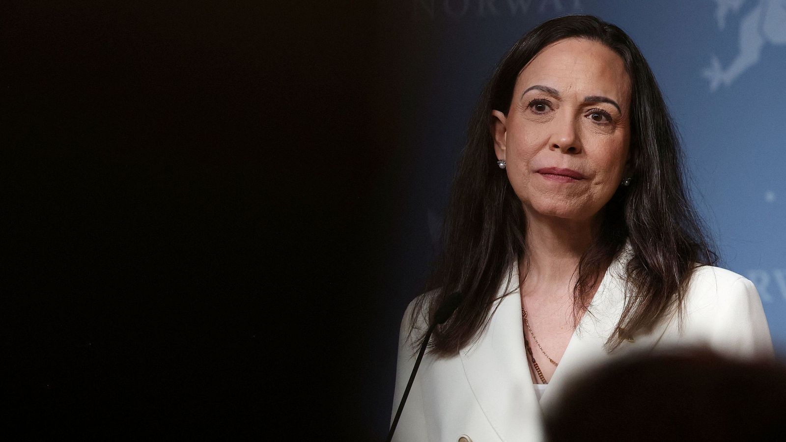 María Corina Machado, con chaqueta blanca y cadena dorada, ofrece una conferencia de prensa. El fondo es azul con un logo.