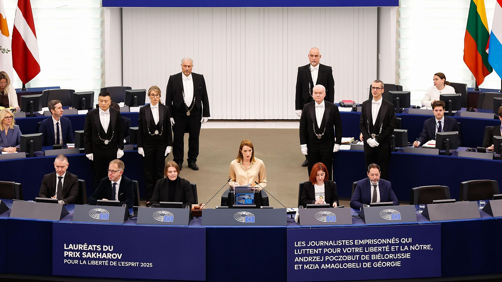 Roberta Metsola preside una ceremonia en el Parlamento Europeo, con un micrófono frente a ella. Se observa a personas vestidas formalmente y banderas en el fondo, con textos sobre el Premio Sájarov.