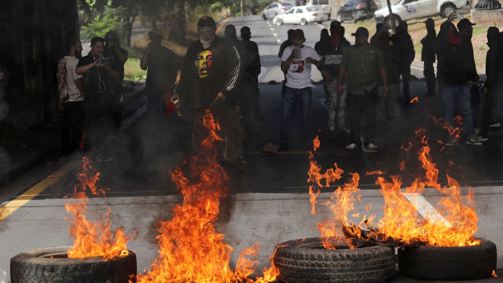 Una protesta en Honduras muestra una barricada de neumáticos en llamas en la calle, con manifestantes observando y grabando la escena. Se observa a un manifestante con una sudadera negra y al fondo vehículos, incluyendo un coche policial, en un ambiente de tensión.