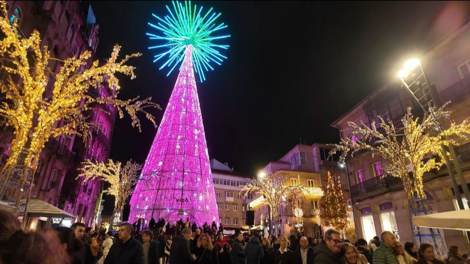 Una plaza en una ciudad española se ilumina con luces navideñas, destacando un gran árbol cónico con luces rosas y una estructura superior verde azulada. La escena está llena de gente disfrutando del ambiente festivo.