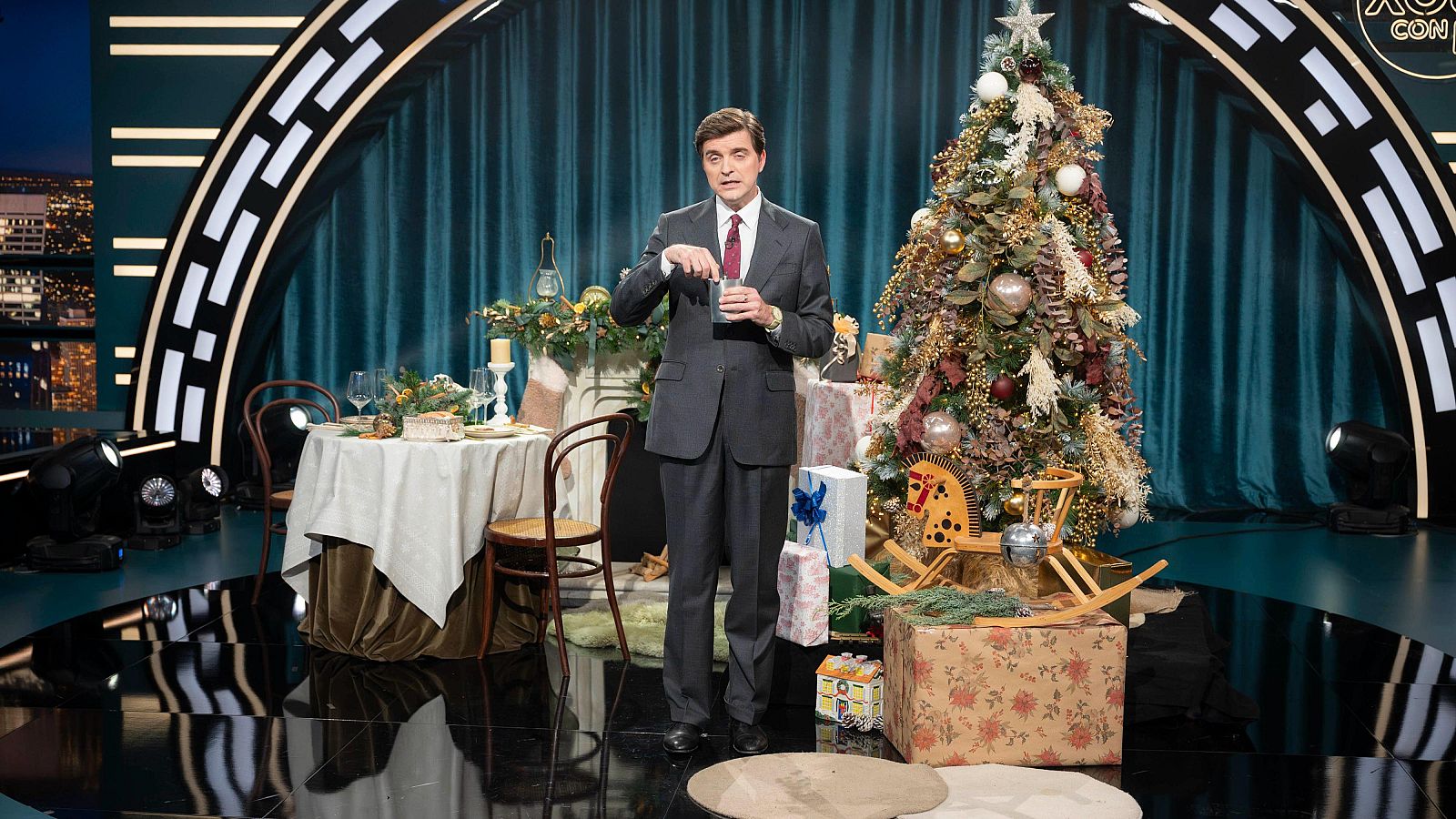 En un escenario decorado para la Navidad, un hombre vestido con traje y corbata sostiene un vaso. A su alrededor se observan elementos festivos como un árbol de Navidad y una mesa con adornos, con un fondo de cortinas y luces.