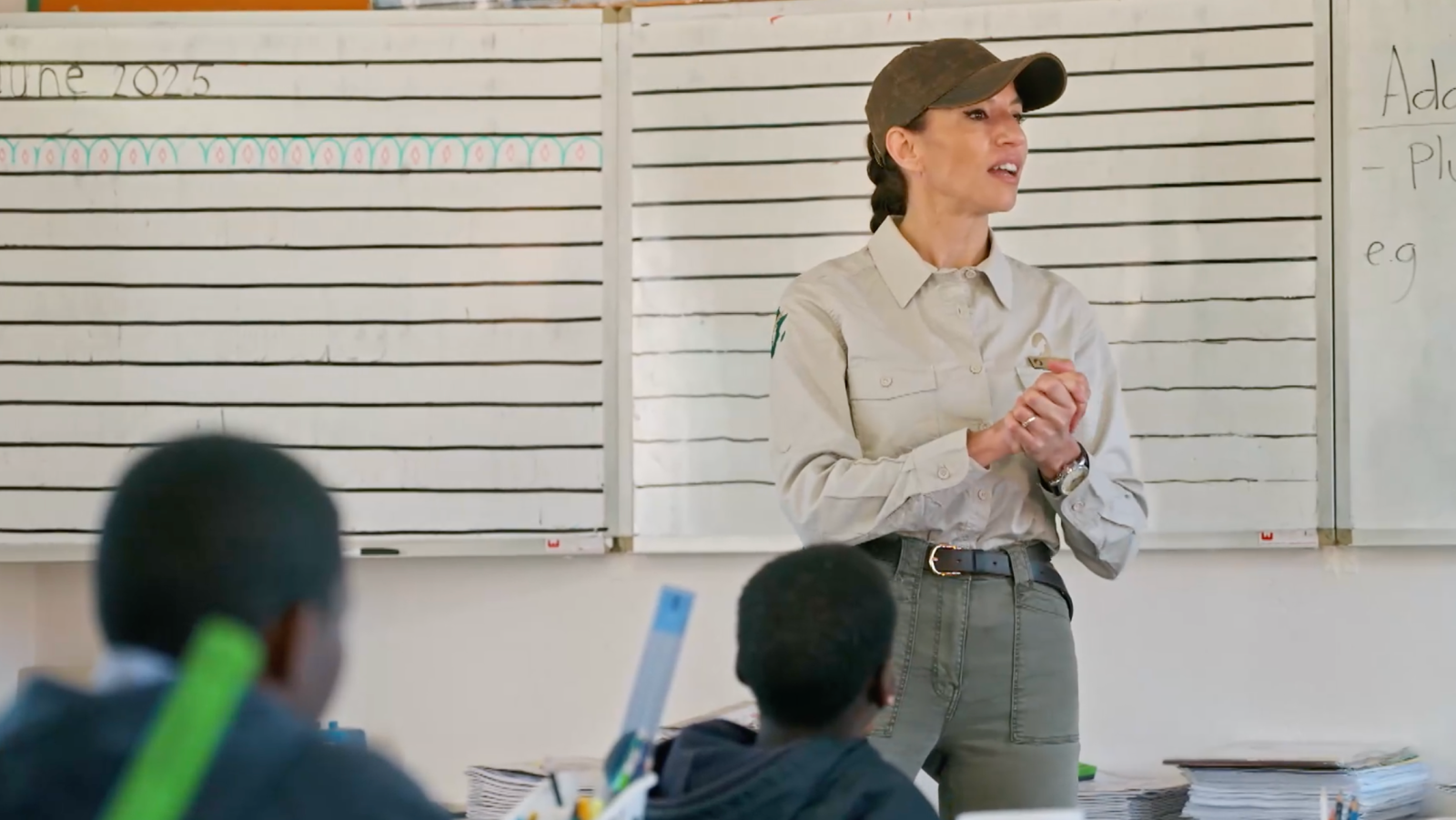 En un aula escolar de Namibia, la Dra. Jones, con gorra y vestimenta informal, se dirige a los estudiantes. Detrás de ella, dos pizarras muestran textos y líneas, posiblemente relacionadas con una lección.