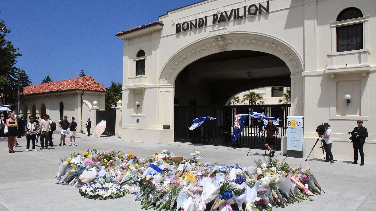En el exterior del Bondi Pavilion, un edificio claro con un arco de entrada, se observa un memorial floral con banderas. Hay personas presentes, algunas con equipos de televisión.