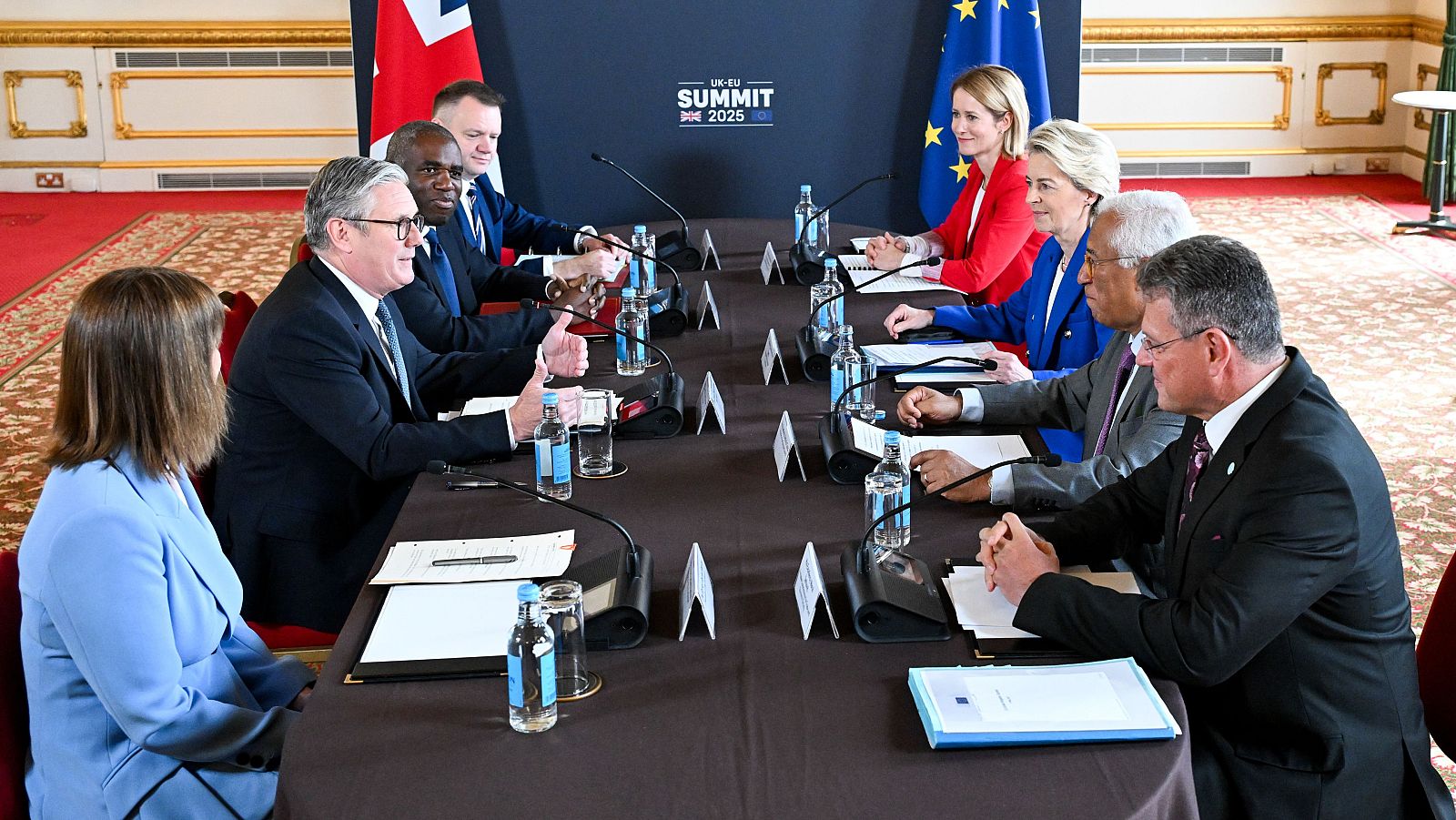 En una mesa de reuniones, se observa a varios individuos, incluyendo a un hombre con traje oscuro y una mujer con chaqueta roja, junto a micrófonos, botellas de agua y documentos. Se distinguen banderas y un cartel con la inscripción "UK-EU SUMMIT 2025".