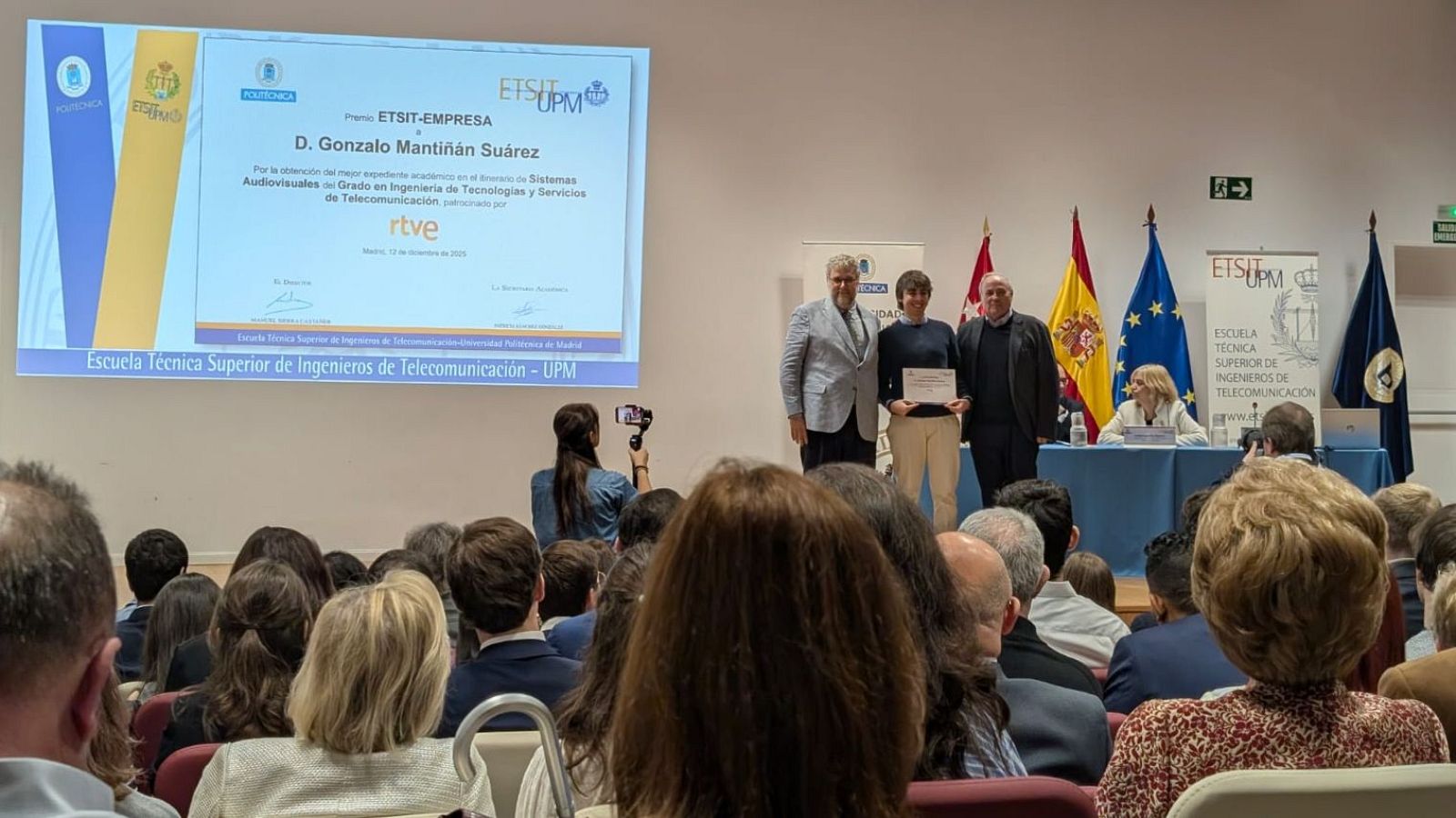 En la ceremonia de entrega de un diploma en la ETSIT-UPM, se observa a tres personas en el escenario. El diploma, con el logo de la institución, es entregado a un estudiante destacado, con una audiencia presente.