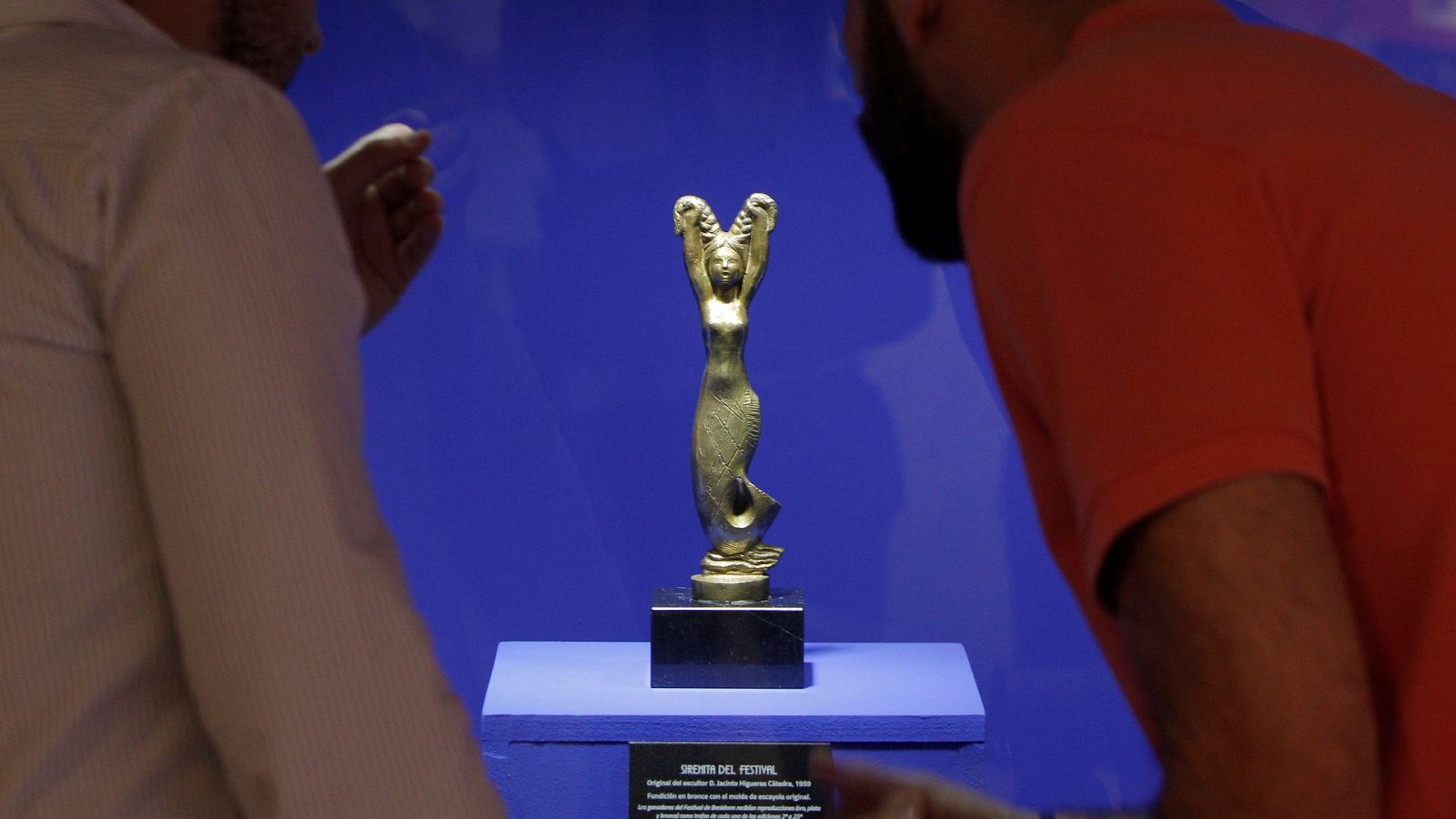 Una estatuilla dorada con forma de sirena, sobre una base negra y plataforma azul, es observada por dos personas. Una de ellas viste una camisa a rayas y la otra una camiseta naranja con mascarilla.