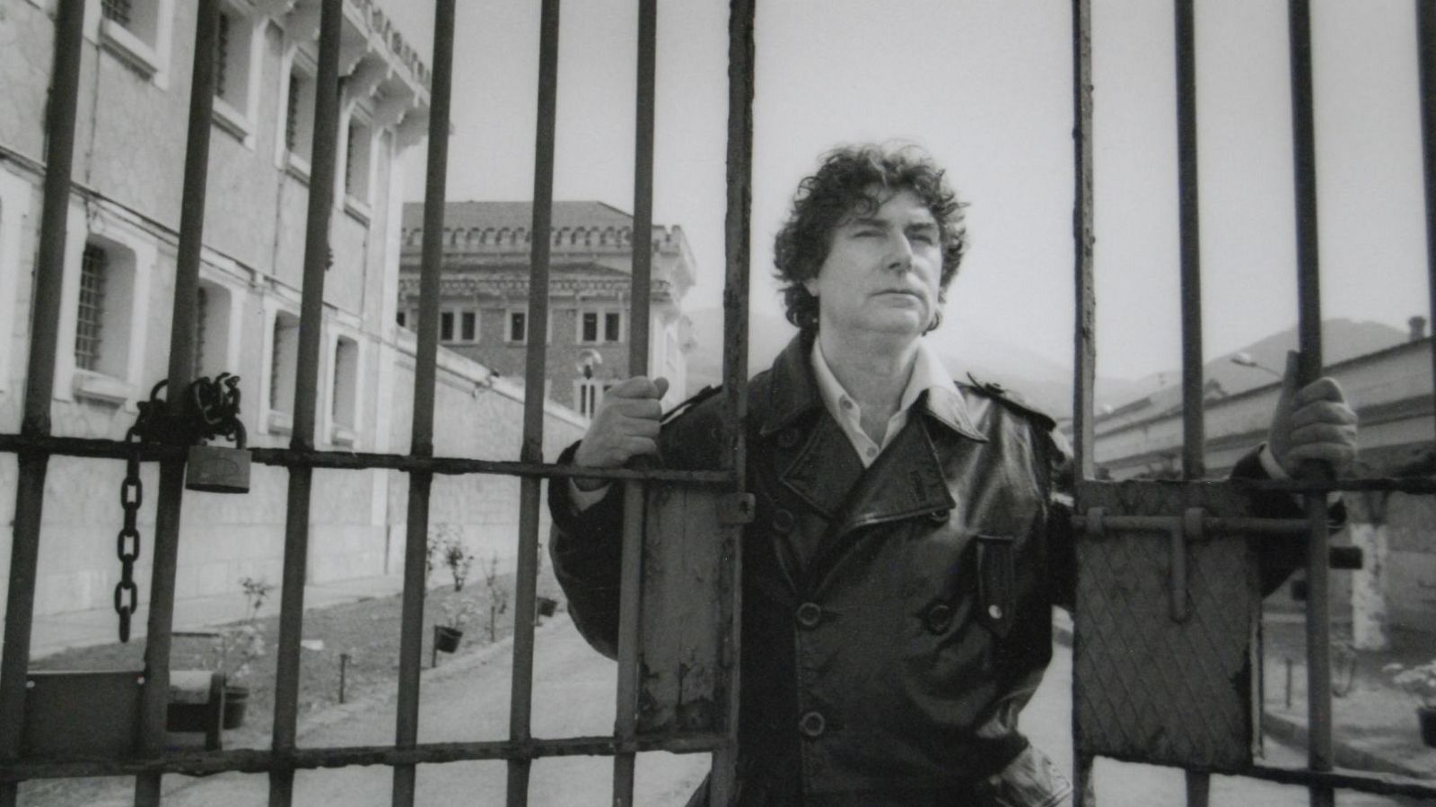 Jesús Quintero, con chaqueta de cuero y camisa clara, se encuentra tras una reja. Su mirada se dirige hacia la derecha, con expresión pensativa, mientras sus manos sostienen los barrotes.