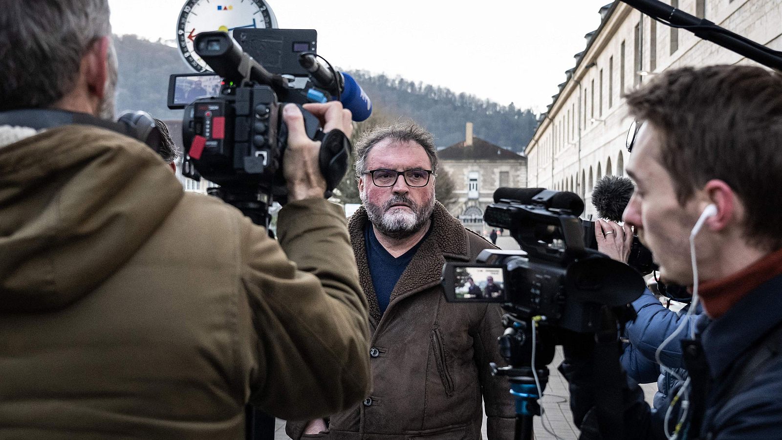 Frederic Pechier, con gafas y barba, es captado por las cámaras de los periodistas. Viste una chaqueta marrón y una camiseta azul, mientras se encuentra frente a un edificio con numerosas ventanas.