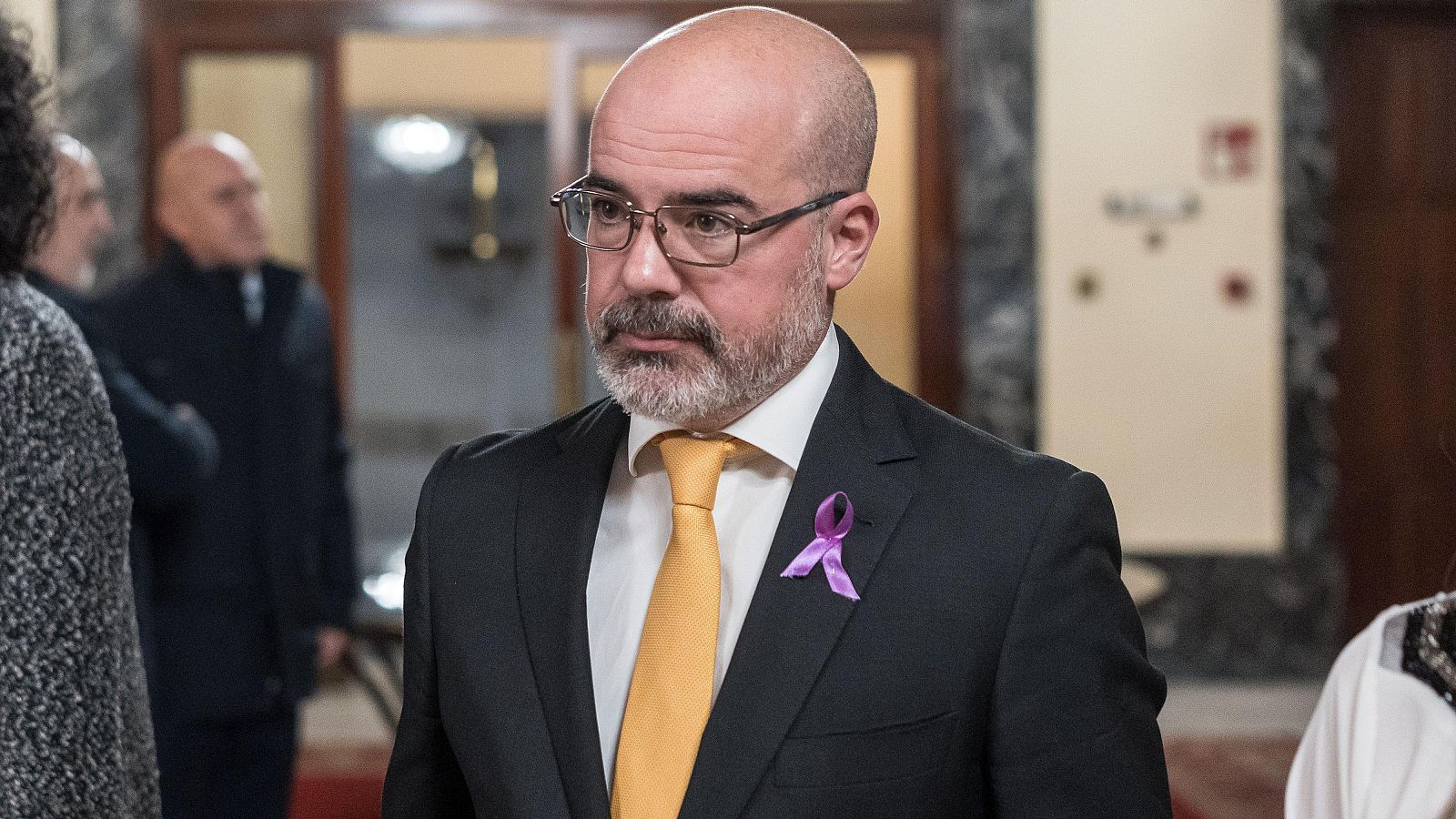 Un hombre mayor, con gafas y barba gris, ataviado con traje oscuro, camisa blanca, corbata amarilla y un lazo morado, posa en un interior.