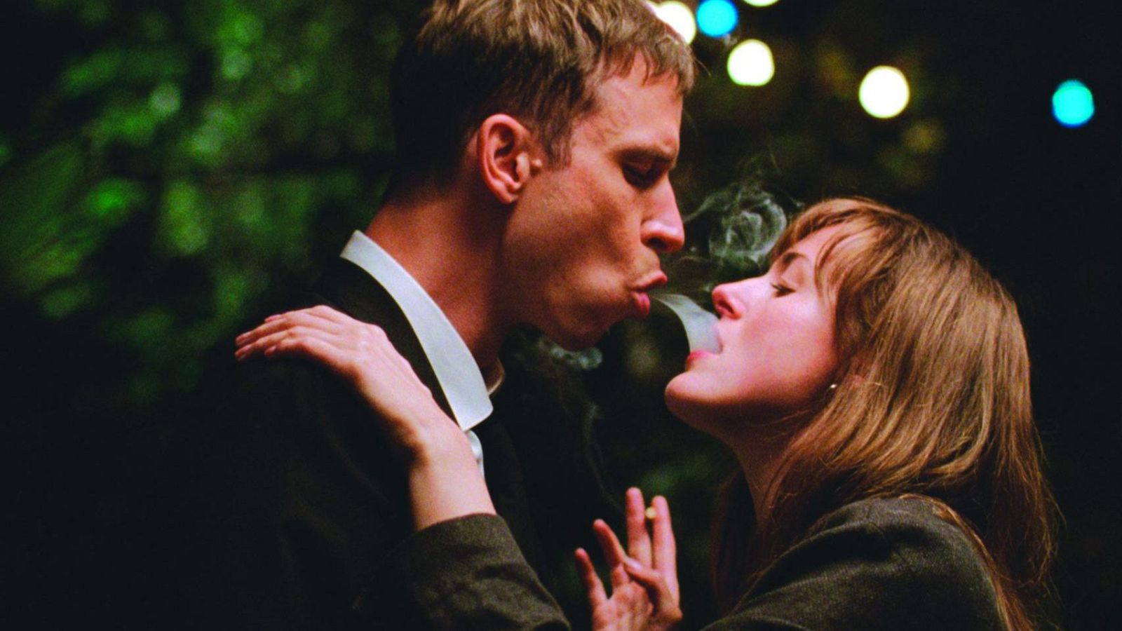 Una fotografía captura a Herbert Nordrum y Renate Reinsve, posiblemente en una escena de película. Los actores, muy cercanos, exhalan humo en un ambiente oscuro con luces borrosas, creando una atmósfera íntima.