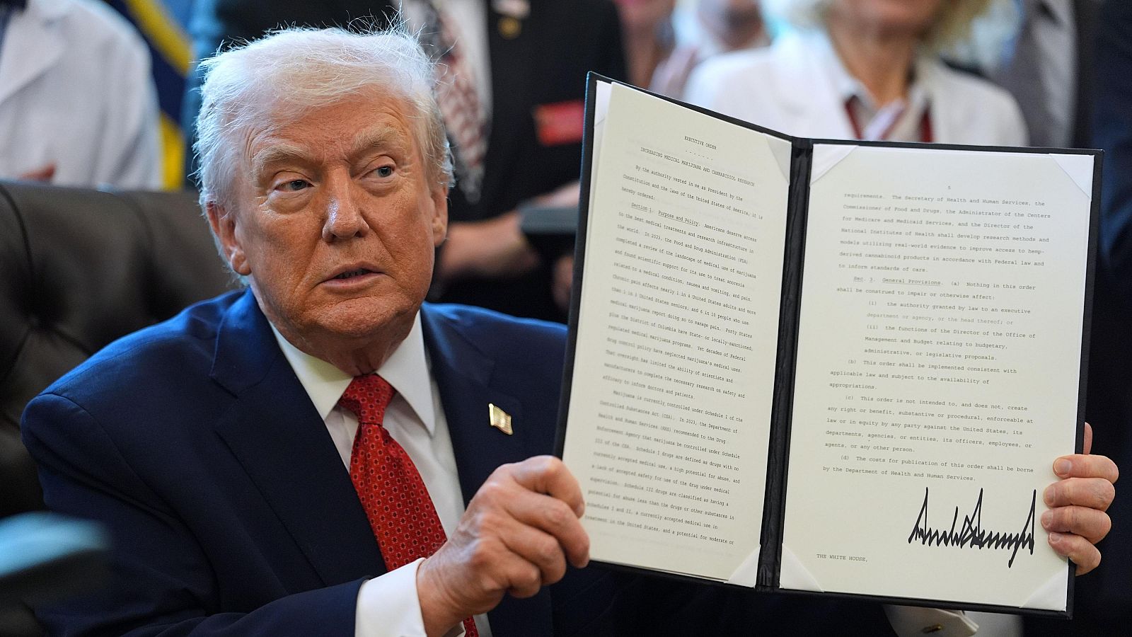 Un hombre con traje azul y corbata roja sostiene un documento con texto y una firma, posiblemente una orden o ley, mientras otras personas se encuentran en el fondo.