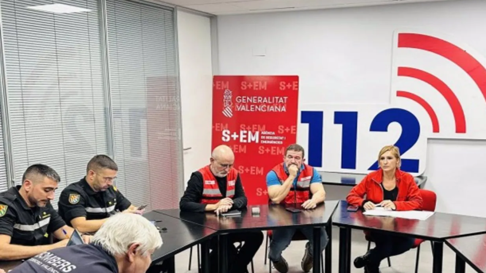 En una sala de reuniones, un grupo de personas, incluyendo bomberos y personal de emergencias, están sentados alrededor de mesas. Al fondo, un cartel con el logo de una institución y el número 112 sugieren una reunión relacionada con emergencias.
