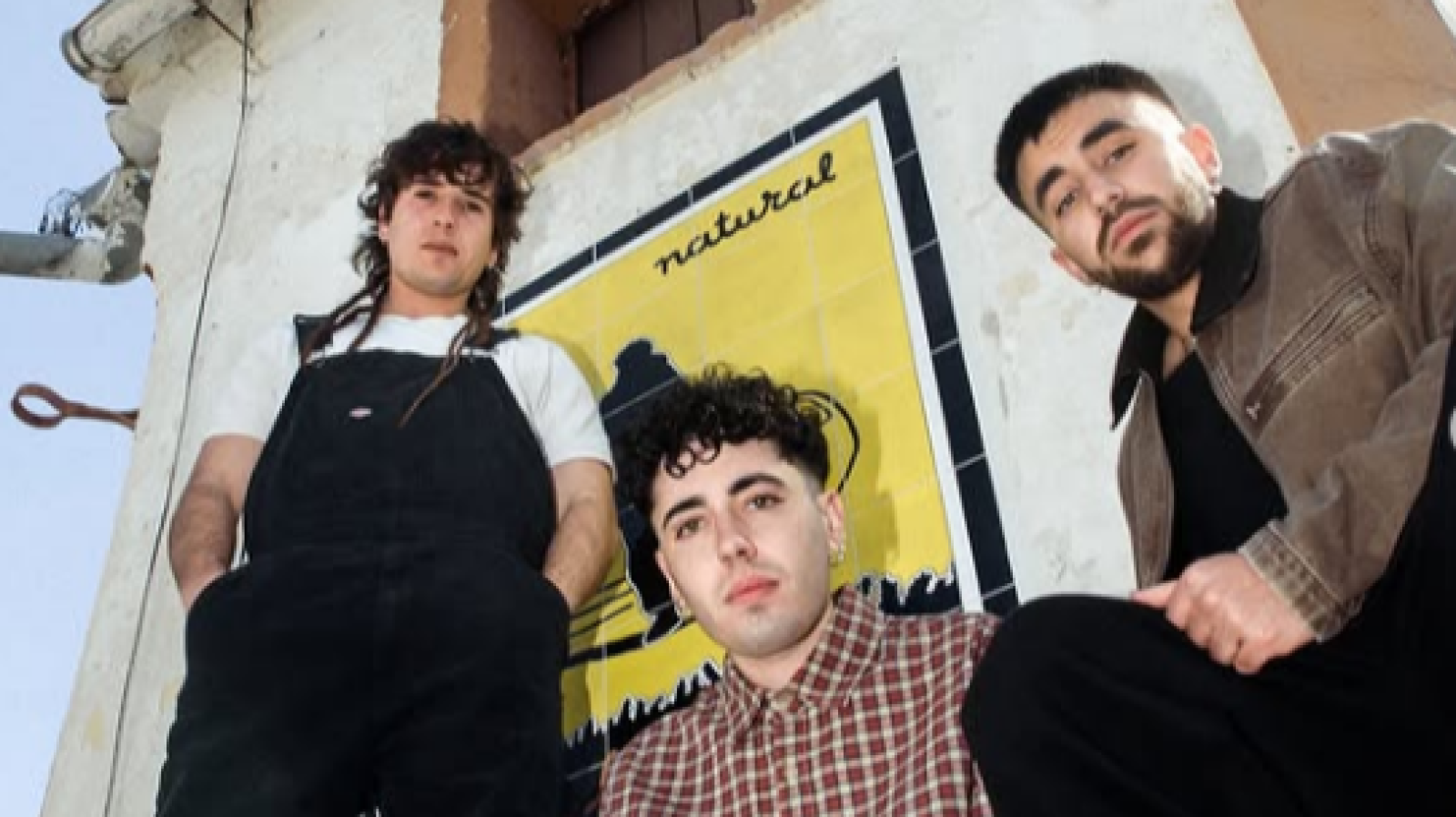 Tres jóvenes se encuentran frente a una pared con azulejos decorativos que muestran la palabra "natural". Uno lleva un mono negro, otro una camisa de cuadros y el tercero una chaqueta marrón.