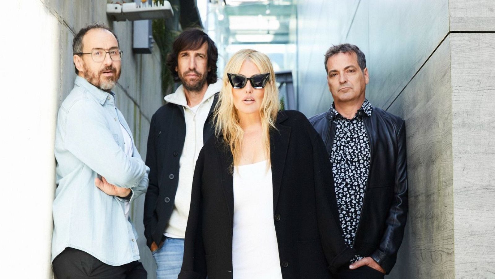 Fotografía de los integrantes de la banda posando en un ambiente con paredes claras. Destaca una mujer con gafas de sol y un vestido blanco, acompañada por tres hombres con diferentes estilos de vestimenta.