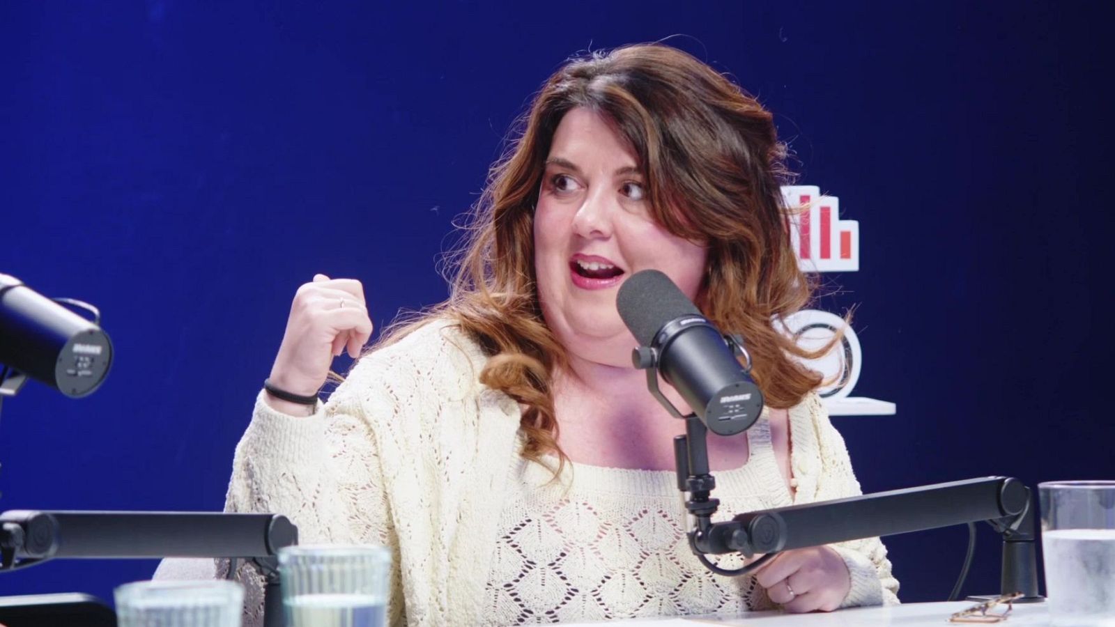Lalachus, con un jersey crema, está frente a un micrófono Shure en un estudio de radio, posiblemente grabando un podcast, gesticulando con la mano izquierda. En la mesa hay vasos y micrófonos, con un fondo azul oscuro y un logo de "Radio".