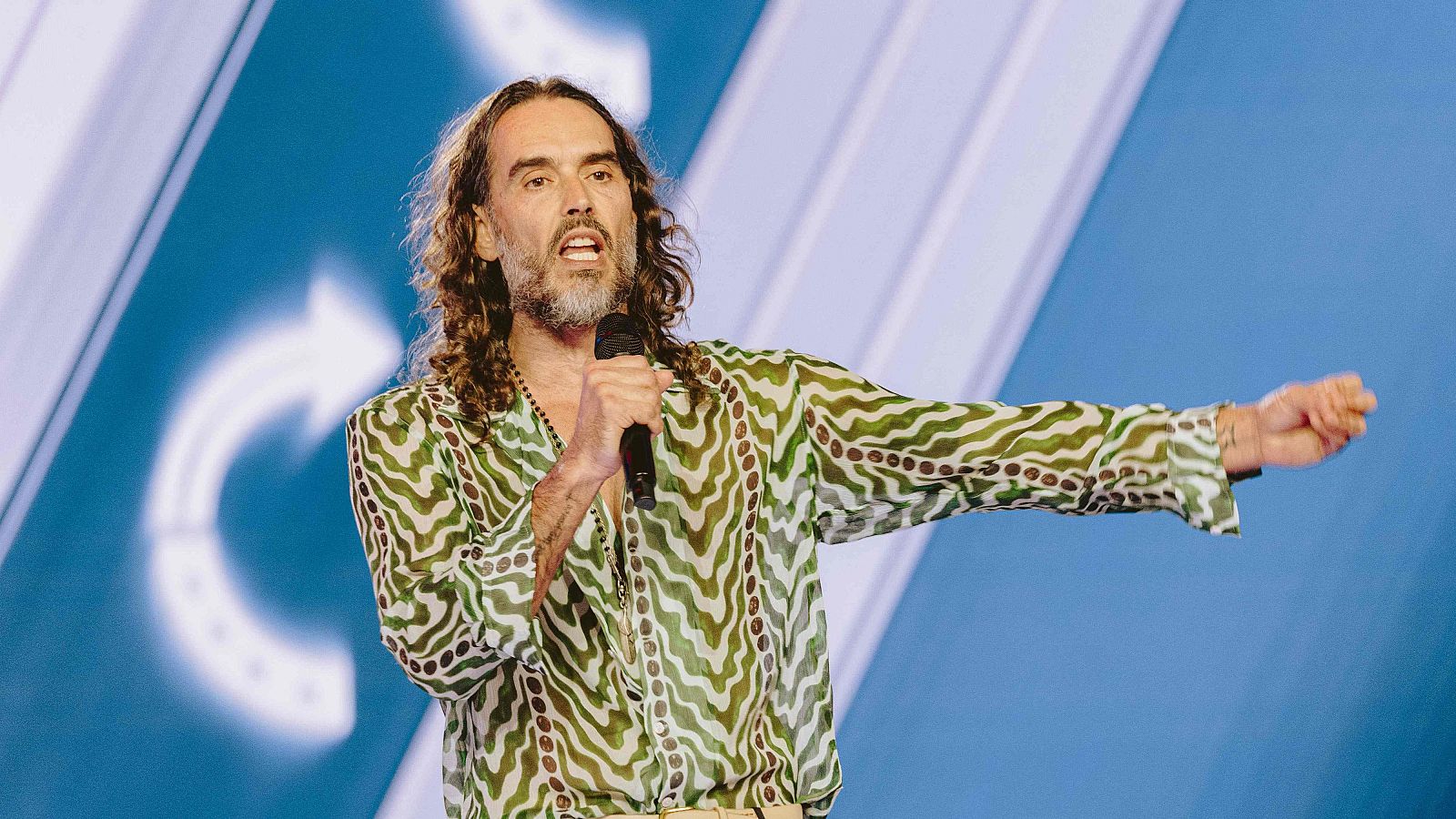 Russell Brand, con su característico cabello largo y barba, actúa en un escenario. Viste una camisa estampada y un collar, sosteniendo un micrófono y gesticulando con la mano.