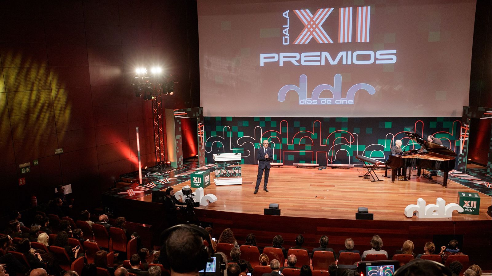 Un presentador en traje oscuro se encuentra en el escenario de una gala, junto a un pianista que toca un piano de cola. El fondo muestra el logo de un programa y el título del evento, con una audiencia numerosa frente al escenario.