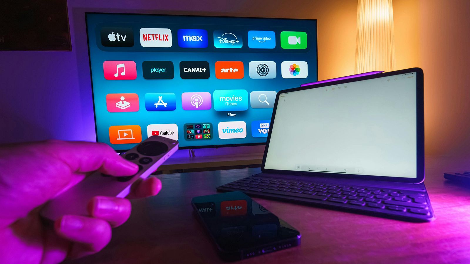Un televisor muestra iconos de aplicaciones de streaming. En la mesa, hay una tableta con teclado y un teléfono móvil. Una mano sostiene un mando a distancia.