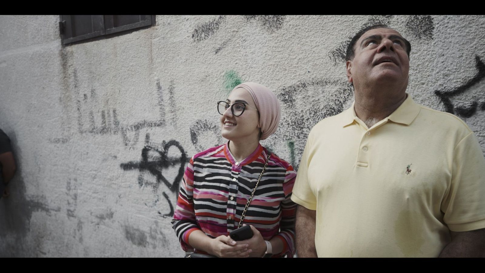Una joven con gafas y pañuelo rosa, sonríe mientras sostiene un teléfono, acompañada por un hombre de mediana edad con un polo amarillo, ambos frente a una pared con grafitis.