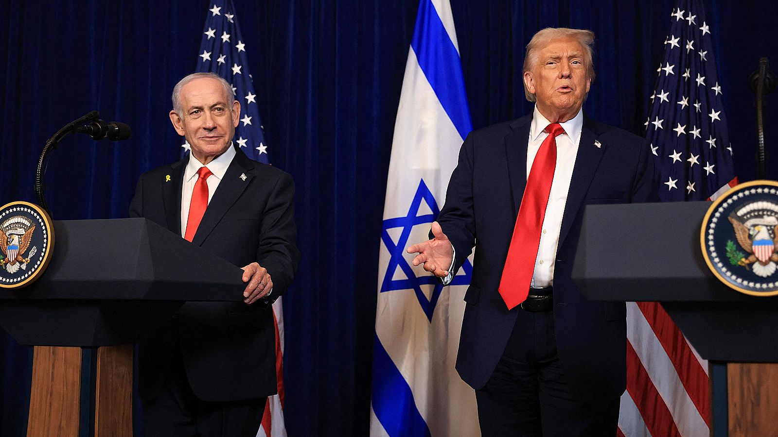 Donald Trump y Benjamin Netanyahu se saludan con un apretón de manos, ambos vestidos con trajes oscuros y corbatas rojas, sonriendo frente a una puerta ornamentada y una bandera.