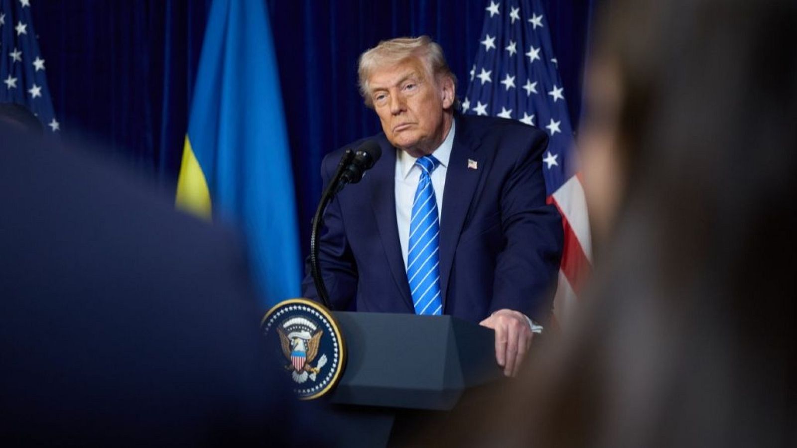 Donald Trump, con traje oscuro y corbata, habla desde un atril con el sello presidencial, con las banderas de Estados Unidos y Ucrania de fondo.
