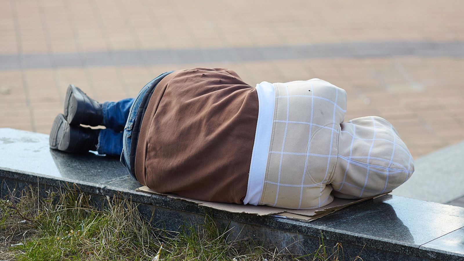 Una persona descansa sobre un banco de piedra, usando ropa casual como pantalones vaqueros y una sudadera. El individuo está acostado sobre un cartón, en un entorno con césped.