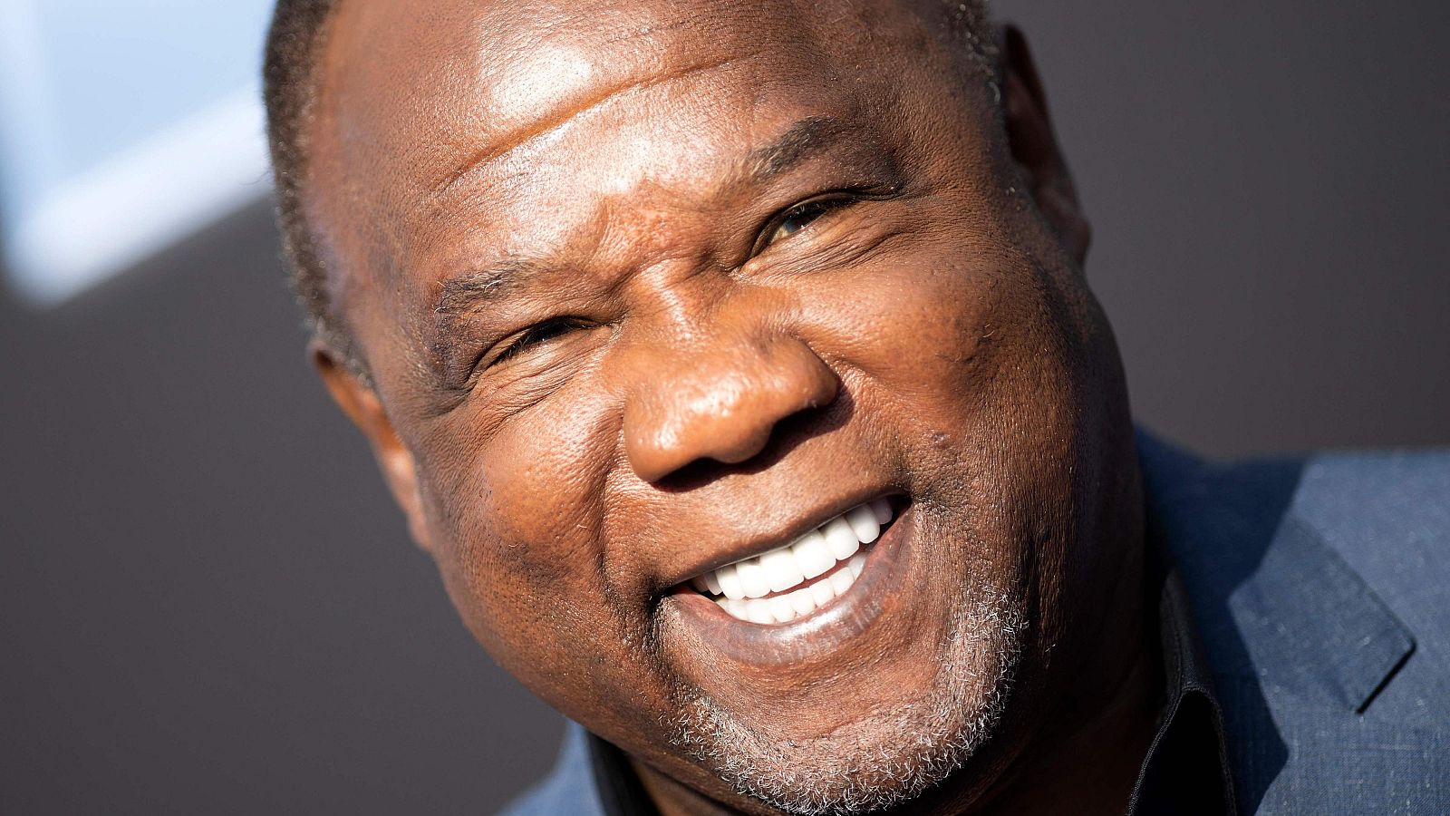 Un hombre afroamericano de mediana edad, con una sonrisa amplia y canas en la barba, posa para la cámara. Lleva una chaqueta azul y su rostro, con arrugas, está bien iluminado.