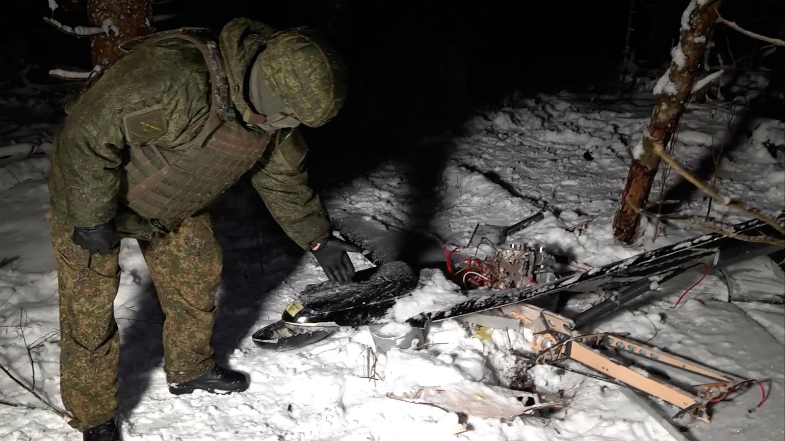 Un militar, vestido con uniforme de camuflaje y guantes negros, inspecciona los restos de un dron destrozado sobre la nieve. El dron, desmembrado, muestra componentes electrónicos y partes de su estructura en un entorno boscoso y nevado.