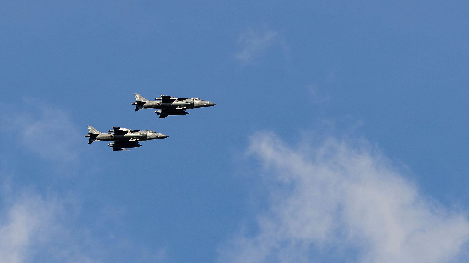 Dos aviones de combate grises vuelan en formación sobre un cielo azul con nubes blancas, uno ligeramente por delante del otro, sugiriendo una exhibición aérea.