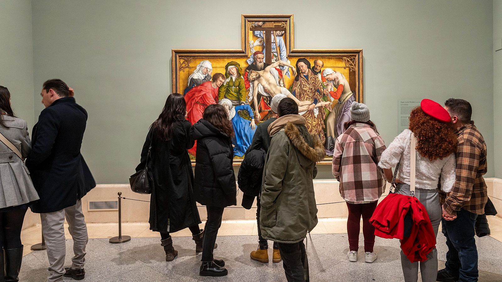 En el Museo del Prado, un grupo de personas observa una pintura religiosa, un tríptico dorado que representa una escena religiosa. Se distinguen varias personas de espaldas, algunas con abrigos y otras con camisas, absortas en la contemplación de la obra.