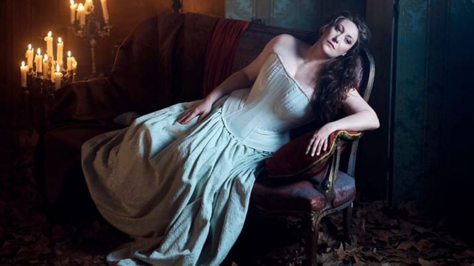 Una mujer con vestido claro y corsé, con el cabello suelto, se recuesta en un sofá oscuro. La escena, con un candelabro y cortinajes rojos, evoca una atmósfera dramática y otoñal.