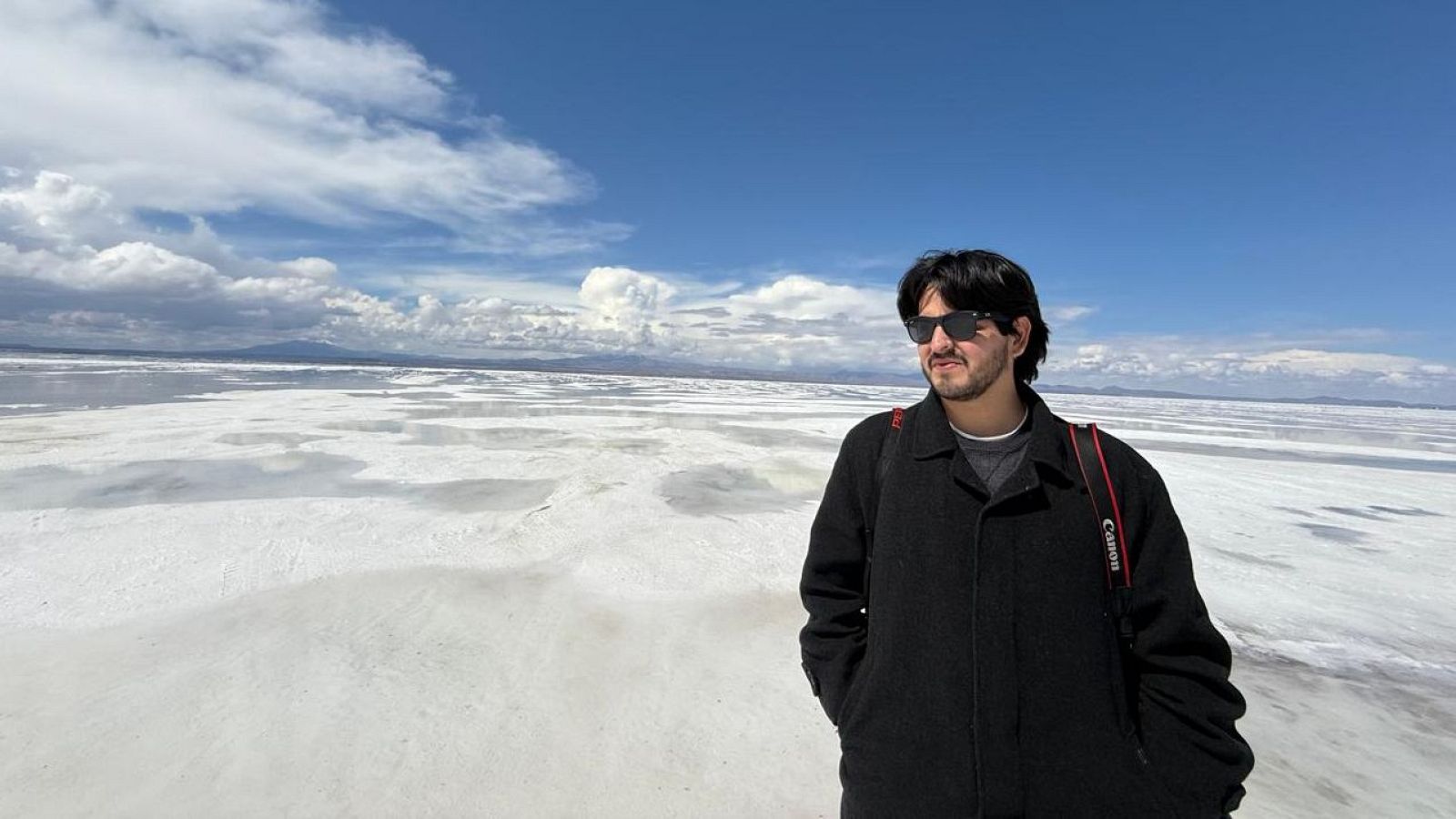 Un hombre con gafas de sol y chaqueta oscura, posiblemente un periodista, posa con una cámara fotográfica. El fondo es un paisaje extenso y blanco, con un cielo azul y nubes.